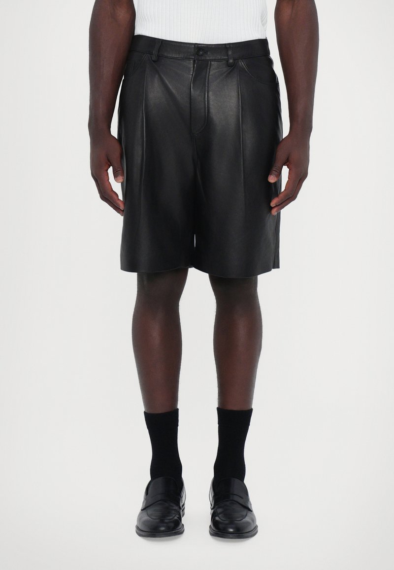 Shorts en cuir noir avec des plis, taille ajustée et texture lisse. Porté avec des chaussettes noires et des mocassins, présenté sur un fond neutre.