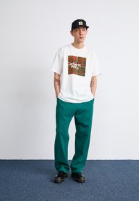 Biela grafická T-shirt s farebným kvetinovým dizajnom a textom "Carhartt WIP", spárovaná s tyrkysovými nohavicami a čiernymi topánkami.