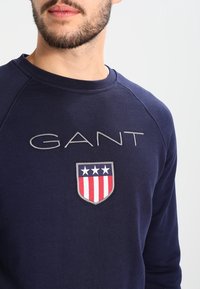 Marineblauwe sweatshirt met "GANT"-tekst en een schildlogo met drie sterren en rode strepen. Gemaakt van zacht materiaal met een ronde halsontwerp.