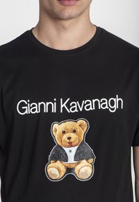 Čierne bavlnené tričko s potlačou medveďa v čiernej bunde a textom "Gianni Kavanagh" bielou farbou pod designom.