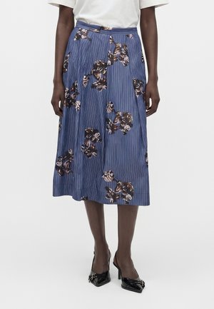 Femme portant une jupe bleue longueur genou avec des rayures verticales et des motifs floraux marron, associée à des talons pointus noirs et un haut blanc.
