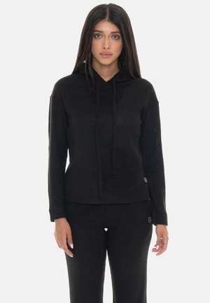 Vrouw met lang donker haar die een zwarte gebreide hoodie en bijpassende zwarte gebreide broek draagt tegen een effen achtergrond.