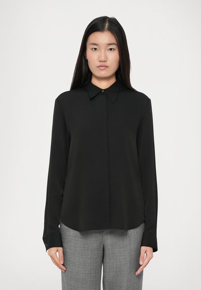 Femme portant une blouse noire à manches longues avec un col et un pantalon à carreaux gris, se tenant devant un fond blanc uni.