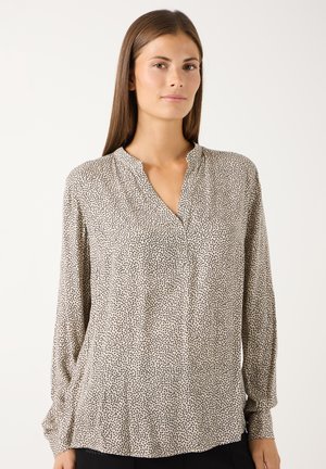 Frau mit glatten braunen Haaren, die eine lockere beige Bluse mit kleinen schwarzen Punkten und V-Ausschnitt trägt, vor einem einfarbigen Hintergrund stehend.