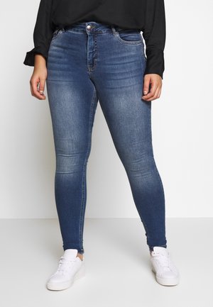 Osoba ubrana w niebieskie obcisłe dżinsy z delikatnym przetarciem i białe trampki, stoi na jednolitym jasnym tle.