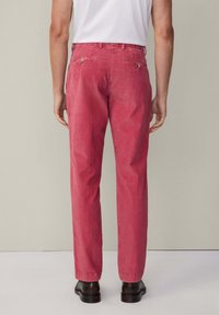 Pantalon en velours côtelé rose avec une jambe droite, deux poches arrière et des accents de boutons métalliques. Porté avec un t-shirt blanc et des chaussures noires.