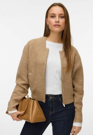 Brauner Strickcardigan mit Reißverschluss, gerippten Bündchen und verkürztem Design, kombiniert mit einem weißen Hemd und einer hellbraunen Handtasche.