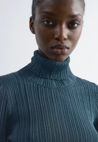 Junge Person mit dunkler Haut, die einen gerippten, hochgeschlossenen, dunkelblauen Pullover vor einem schlichten hellen Hintergrund trägt.