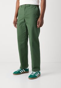 Pantaloni in twill di cotone verde con taglio dritto, tasche laterali e chiusura con un bottone in vita. Abbinati a sneaker verdi.