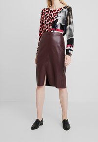 Jupe crayon en cuir bordeaux avec une fente latérale, associée à un haut à manches longues à motifs multicolores. Des chaussures noires faciles à enfiler complètent la tenue.