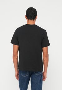 Vyras su trumpais tamsiais plaukais, dėvintis paprastą juodą trumpomis rankovėmis marškinėlį ir mėlynas džinsines kelnes, stovintis nugara į baltą foną.