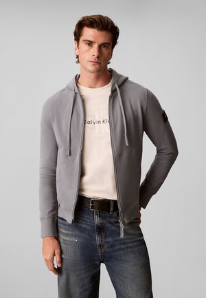 Młody mężczyzna w jasnoszarej bluzie na suwak, na białej koszulce Calvin Klein i niebieskich jeansach, stojący na jednolitym tle.