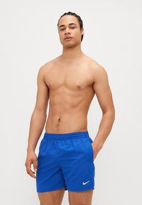 Blå Nike svømmebukser med elastisk talje og sidelommer, har en subtil glans og en hvid logo på nederste venstre side.