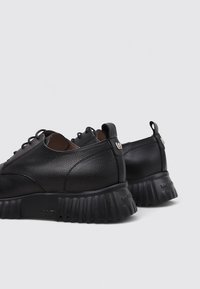 Par de zapatos de cuero negro texturizado con cordones y suelas gruesas y estriadas, vistos desde atrás sobre un fondo gris claro.