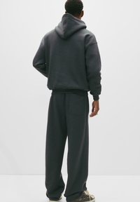 Person som bär en mörk oversized hoodie och matchande vida byxor, stående med ryggen vänd mot kameran mot en enkel bakgrund.