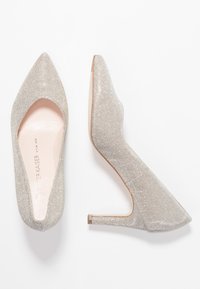 Srebrne szpilki mają spiczaste noski, teksturowany, błyszczący materiał oraz unikalny wzór z wycięciem na pięcie. Gładkie wewnętrzne wykończenie.