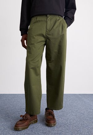 Mężczyzna noszący oliwkowe szerokie spodnie, czarną bluzkę z długim rękawem oraz brązowe mokasyny z frędzlami, stojący na szarym dywanie.