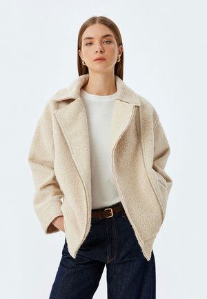 Beige uldblandet frakke med en notch krave, oversized pasform og diagonal lynlåsåbning. Kombineret med en hvid top og mørk denimjeans.