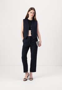 Námornícky pruhovaný vest s gombíkmi, doplnený o zladene nohavice a čierny crop top. Drží textúrovanú kabelku a má na sebe remienkové sandále.