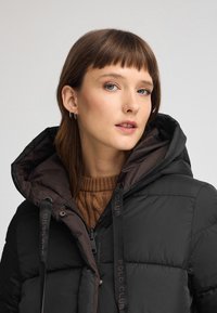 Czarna kurtka puchowa z brązowym kapturem, z przeszytymi panelami, zamkiem błyskawicznym i markowymi sznurkami do ściągania. Noszona na brązowym swetrze dzianinowym.