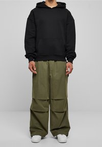 Sudadera negra con capucha y bolsillo frontal, combinada con pantalones cargo sueltos de color verde oliva con múltiples pliegues. Zapatillas blancas completan el look.