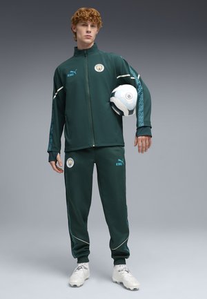 Jeune homme en survêtement vert foncé de Manchester City tenant un ballon de football blanc et bleu sur un fond gris.