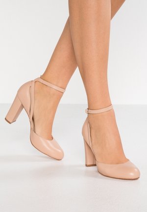 Zapatos de tacón alto beige con puntera redondeada, correa en el tobillo y tacón cuadrado afilado. Acabado de cuero suave con diseño de cortes a los lados.