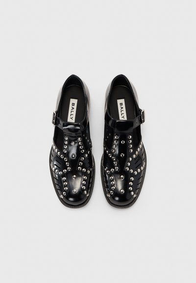 Sapatos pretos brilhantes com uma tira, apresentando tachas prateadas, padrões recortados e uma sola texturizada. Logotipo visível na palmilha.