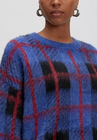 Eine Person trägt einen kuscheligen blauen Pullover mit rot-schwarz kariertem Muster und silbernen Ohrhängern, sichtbar vor neutralem Hintergrund.