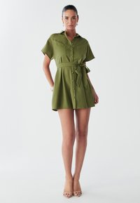Abito camicia verde oliva con maniche corte, apertura frontale con bottoni, cintura in vita e due tasche sul petto. Tessuto morbido e forma ad A.