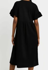 Kvinna i en svart midiklänning med korta volangärmar, sedd bakifrån mot en enkel ljus bakgrund.