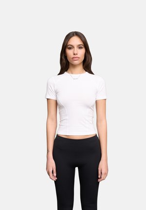 Vrouw staat frontaal, draagt een nauwsluitend wit kortermig shirt en zwarte legging tegen een effen lichtgrijze achtergrond.