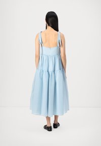 Vestido midi em azul claro, com camadas, feito de tecido suave, com finas alças ajustáveis e fecho de correr nas costas. Sapatos pretos planos.