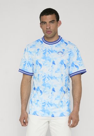 Mężczyzna noszący niebiesko-białą koszulkę w technice tie-dye z paskowanym kołnierzem i rękawami, stojący na jednolitym jasnym tle.