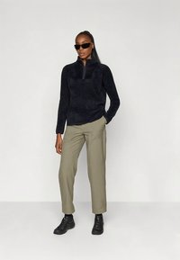 Vrouw met lang haar, gekleed in een zwart fleece-vest, beige broek, zwarte sneakers en een zonnebril, staand tegen een effen lichte achtergrond.