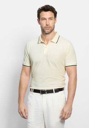 Mann mit kurzen dunklen Haaren und Bart, der ein cremefarbenes Poloshirt mit grünem Besatz und weiße Hose mit schwarzem Gürtel vor weißem Hintergrund trägt.