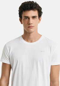 Jonge man met krullend donker haar, gekleed in een eenvoudige witte GANT T-shirt, tegen een effen lichtgrijze achtergrond.