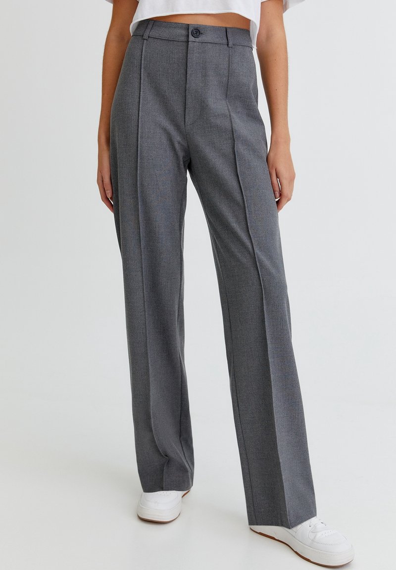 Pantalons gris ajustés à taille haute, avec des plis, une fermeture à crochet et bouton, et une coupe droite. Portés avec des baskets blanches.