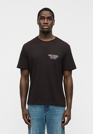 Νεαρός άνδρας με σκούρο δέρμα, ντυμένος με μαύρο μπλουζάκι Michael Kors και μπλε τζιν, στέκεται μπροστά σε απλό ανοιχτό γκρι φόντο.