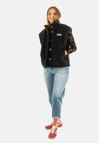 Zwarte, fuzzy fleece vest met opstaand kraag en knoopsluiting. Gecombineerd met licht denim jeans en bordeauxrode sneakers.