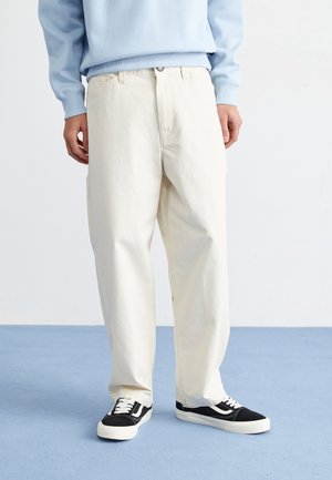 Pantaloni chiari, larghi e in cotone, con taglio dritto e tasche laterali, abbinati a sneaker nere e un maglione blu.