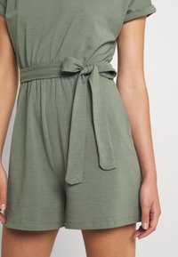 Combinaison courte vert olive avec une ceinture à la taille, col rond, manches courtes et texture lisse. Présente une taille froncée et des poches latérales.