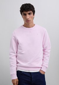 Joven de pie contra un fondo simple, vistiendo un sudadera rosa clara, jeans oscuros y una camisa blanca que asoma por el dobladillo.