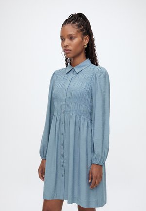 Femme aux cheveux tressés porte une robe bleu clair à manches longues, boutonnée, avec un détail en tissu froncé sur la poitrine et un col pointu.