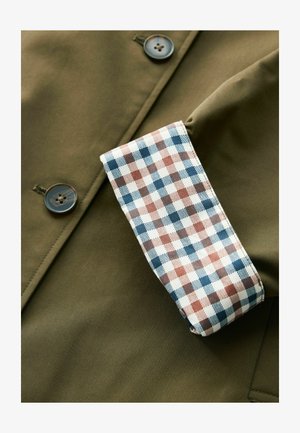 Oliwkowa kurtka o gładkiej fakturze i dwóch guzikach. Rękaw charakteryzuje się białym wzorem w kratkę w odcieniach niebieskiego i brązowego.