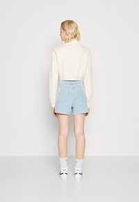 Zastrihnutá svetlá krémová mikina s rebrovaným lemom, doplnená svetlomodrými džínsy šortkami. Model má na sebe biele ponožky a tenisky. Minimalistické pozadie.