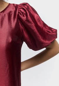 Lesklý hluboce červený saténový top s nafouklým krátkým rukávem a kulatým výstřihem, nošený osobou s jednou rukou ohnutou v pase.