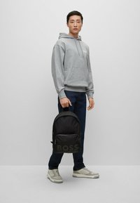 Sweat-shirt gris avec poche avant, jeans bleu foncé, sac à dos noir avec logo "BOSS", et baskets claires, se tenant devant un fond uni.