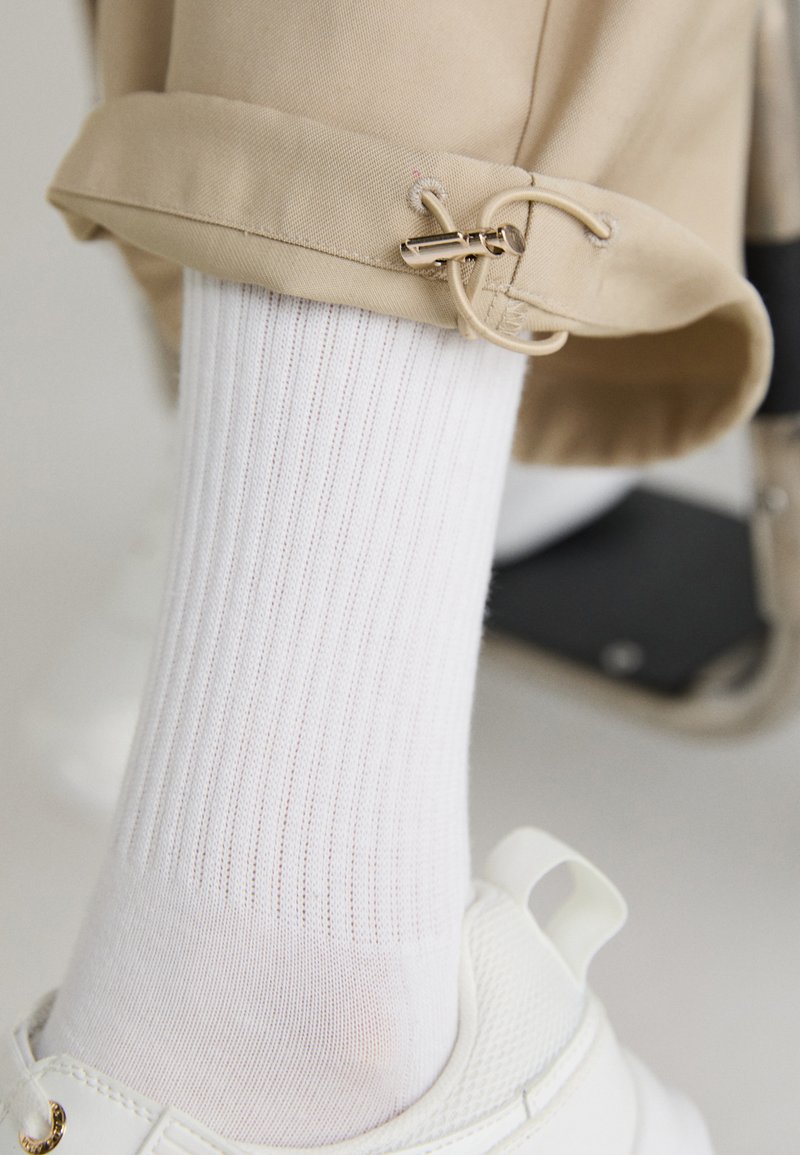Vit ribbade strumpor som är parat med en vit sneaker. Byxan har en beige benvending med en guldfärgad detalj och en ögla-design.