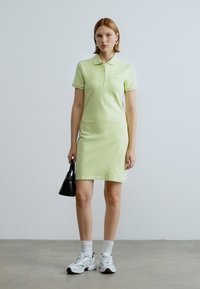 Robe polo vert clair à manches courtes, silhouette ajustée et logo discret. Portée avec des chaussettes blanches et des baskets grises, tenant un sac noir.